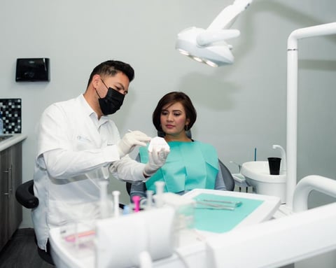 Male dentist wearing white coat and mask examines female patient in turquoise bib in modern dental office with overhead light